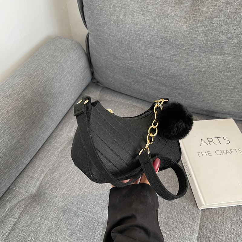 Black handbag with a gold chain and fur pom-pom on a gray couch next to a book titled 'The Arts and The Crafts'.