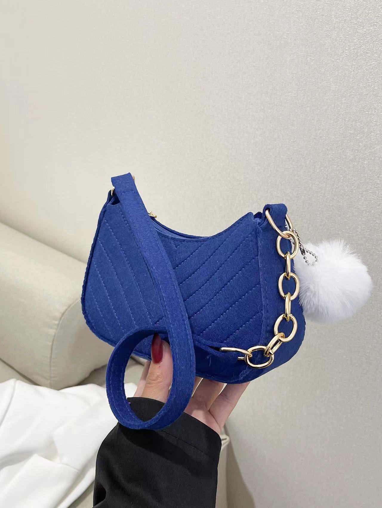 Blue handbag with a chain strap and white pom-pom held by a person against a neutral background