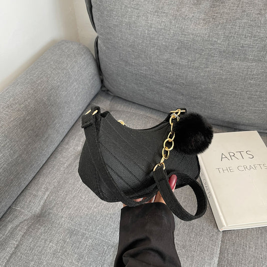 Black handbag with a gold chain and fur pom-pom on a gray couch next to a book titled 'The Arts and The Crafts'.