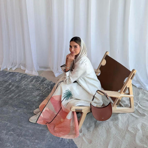 Woman sitting on a chair with a white and pink Modest Floral Style Abaya, holding a brown handbag against a white curtain background.