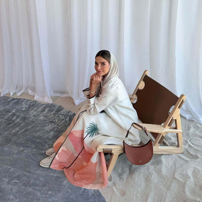 Woman sitting on a chair with a white and pink Modest Floral Style Abaya, holding a brown handbag against a white curtain background.