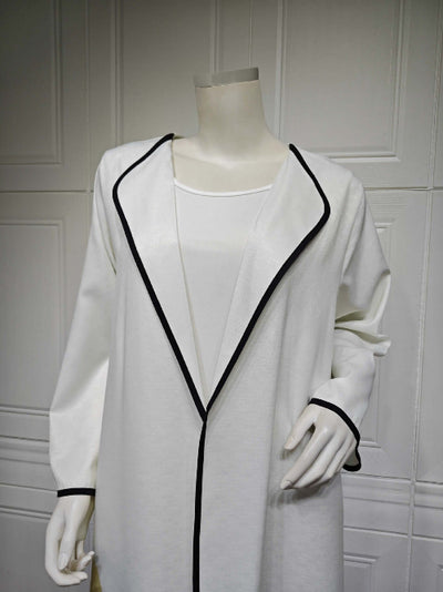 White coat with black trim Modest Floral Style Abaya on a mannequin against a white paneled wall.