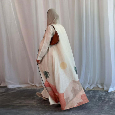 Person wearing a long, patterned Modest Floral Style Abaya with a hood and carrying a brown bag against a white curtain background.