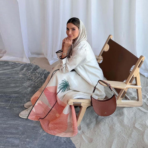 Woman in a white and Modest Floral Style Abaya sitting on a wooden chair with a brown bag.