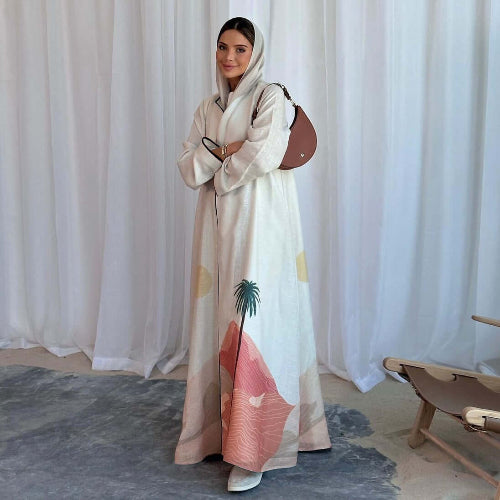 Woman wearing a long, light-colored Modest Floral Style Abaya with a palm tree design against a white curtain background.